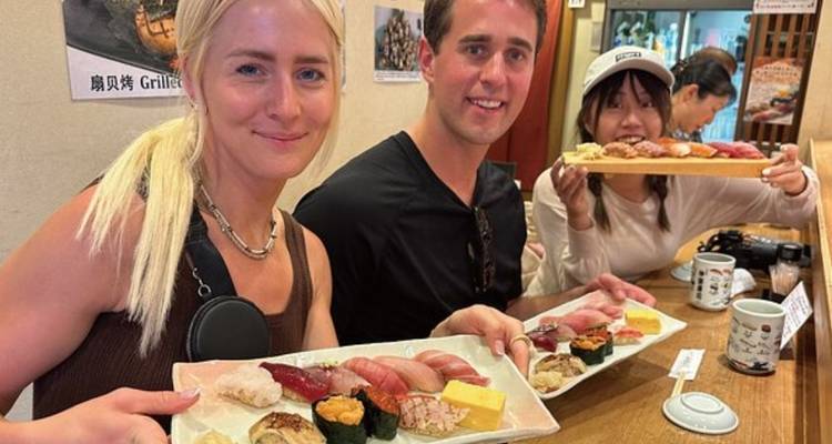 Reisende in einer Sushi-Bar präsentieren Teller mit assortiertem Nigiri und strahlen dabei über das ganze Gesicht.