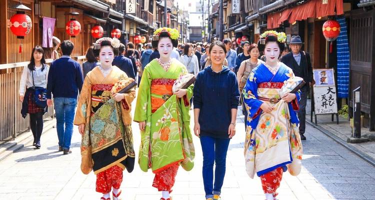 Zwei Maiko und ein Fremdenführer gehen durch das Gion-Viertel von Kyoto, während Zuschauer die traditionelle Kleidung bewundern.