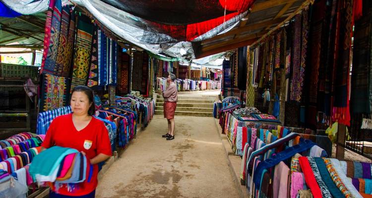 Colourful textile stalls line a covered market alley where shoppers browse fabrics.