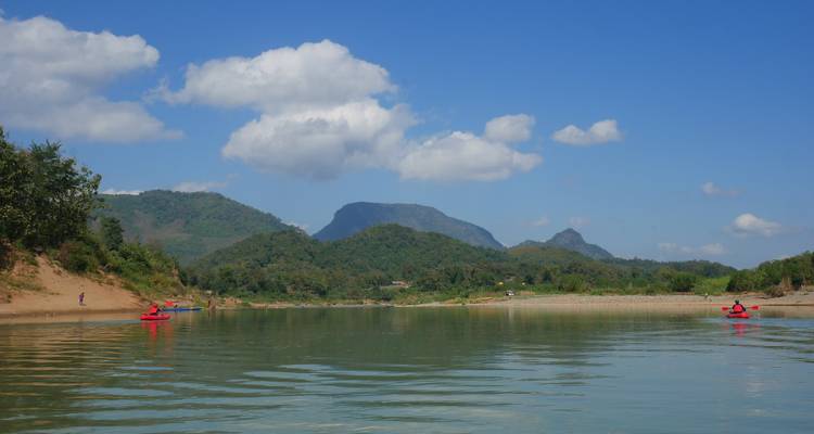 Wide view of tranquil river flanked by forested hills with distant kayakers under scattered clouds.