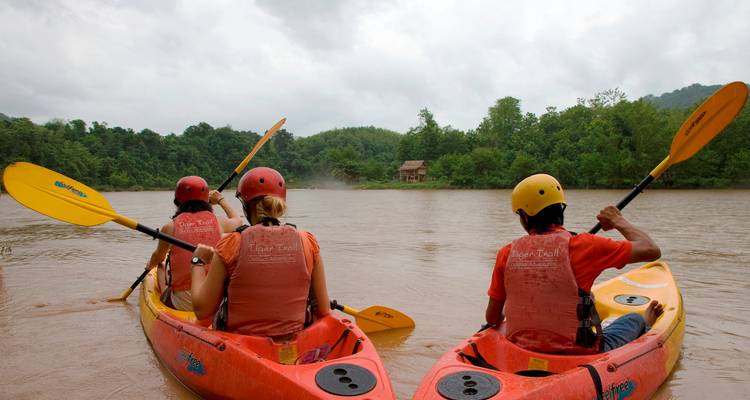 Three kayakers in bright gear sit in their boats on a muddy river, looking out toward a forested bank on an overcast day.