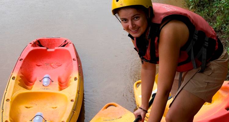 A smiling woman in helmet and life vest steadies her kayak on the riverbank beside brightly colored boats.