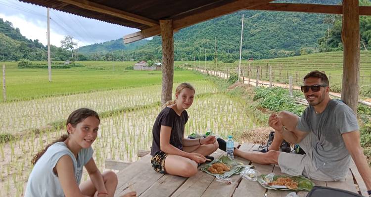 Trois cyclistes font une pause pour un pique-nique sur une plateforme en bois surplombant de jeunes rizières avec des collines verdoyantes en arrière-plan