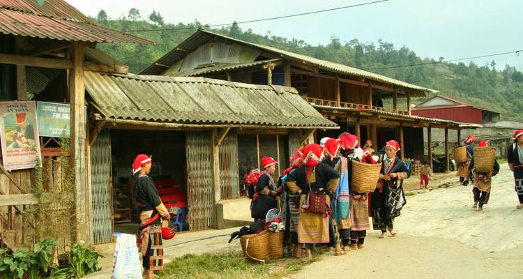 Red-Dao women in traditional dress stand with woven baskets outside rustic wooden shops in a Sapa village.