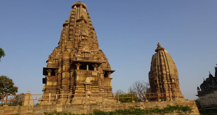 A towering stone spire and a smaller shrine rise against a clear blue sky at a temple complex.