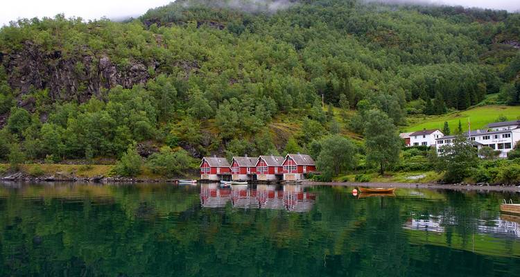Grupo de cabañas rojas en un fiordo verde tranquilo respaldado por una ladera boscosa con neblina baja