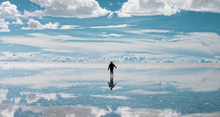 Silhouetted traveler walking across mirror-like Uyuni salt flat reflecting sky and clouds