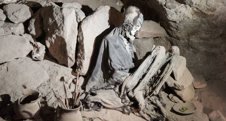 Ancient mummy in seated position inside a rocky cave chamber with artifacts scattered nearby