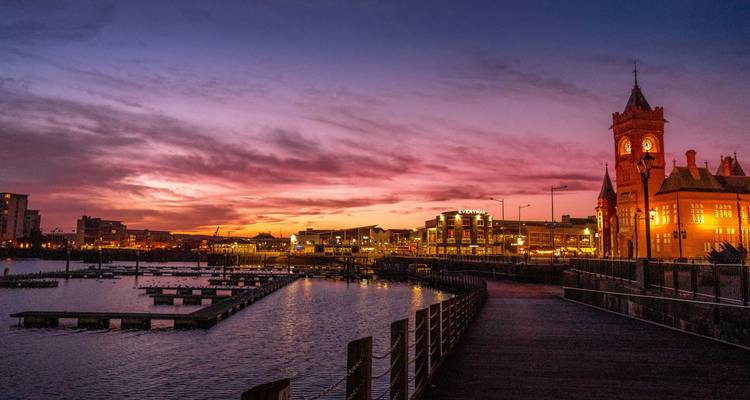 Vista nocturna del puerto deportivo de Cardiff Bay y el histórico edificio Pierhead iluminado bajo un cielo de atardecer púrpura.