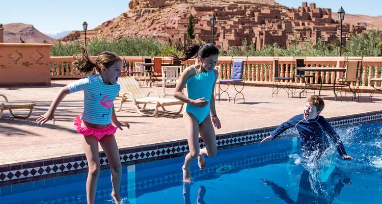 Three excited children leap into a bright blue pool with the adobe kasbah of Ait Benhaddou behind.