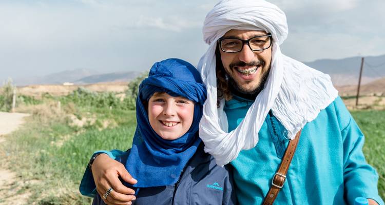 Berber guide and smiling child in traditional turbans pose in rural Moroccan landscape.