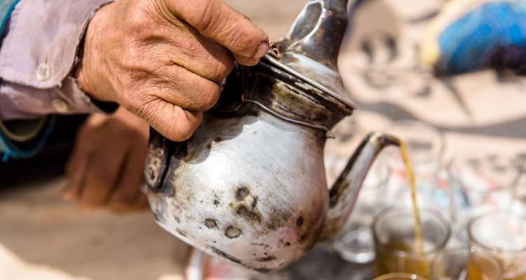Close-up of a weathered hand pouring Moroccan mint tea from a traditional metal pot.