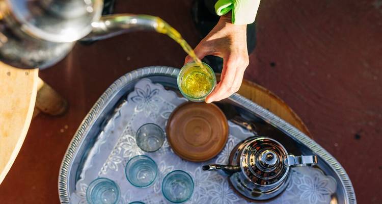Overhead view of golden Moroccan tea being poured into small glasses on a decorative tray.