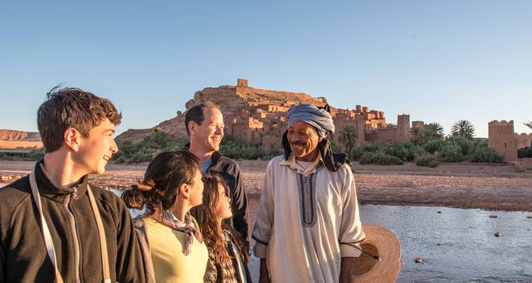 Family chats with local dressed in white robe in front of Ait Benhaddou’s earthen kasbah at sunset.