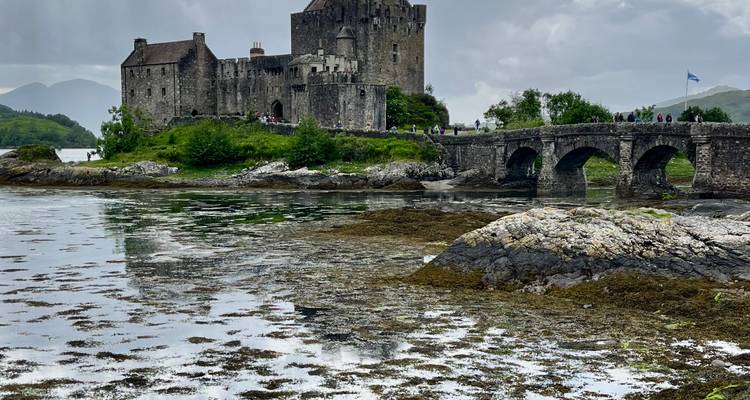 Le château d'Eilean Donan se dresse près d'un pont de pierre surplombant des eaux de marée réfléchissantes et des collines lointaines.