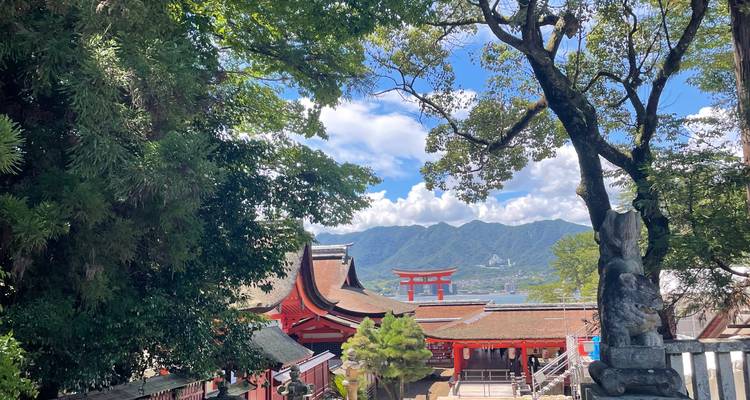 View through lush trees toward the red buildings and iconic floating torii gate of Itsukushima Shrine with mountains beyond.