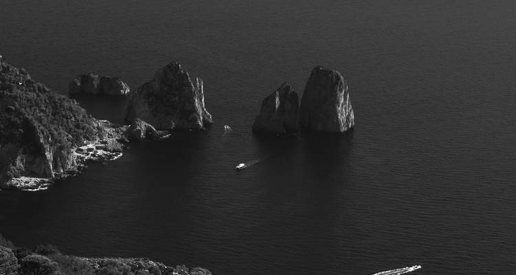 Schwarz-Weiß-Luftaufnahme der Faraglioni-Meeresfelsen vor Capri, umgeben von dunklem Wasser.