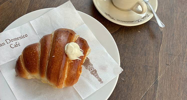 Ein frisches Croissant mit einem Hauch Creme sitzt auf einer Serviette auf einem weißen Teller neben einer Espressotasse auf einem Holzcafétisch.