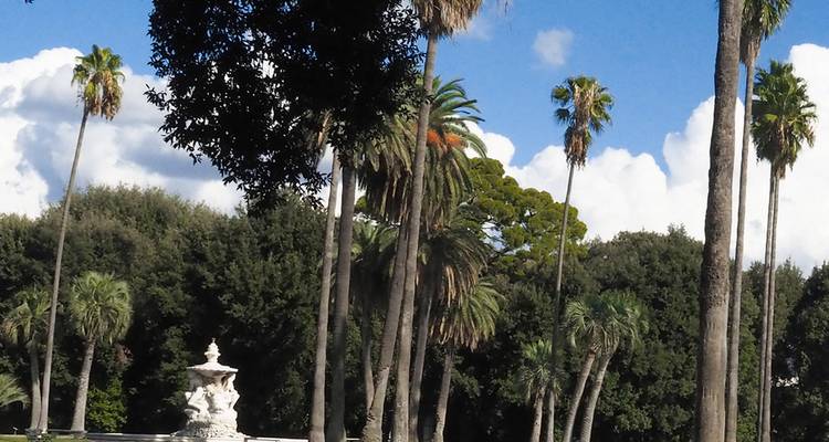 Hohe Palmen ragen über einen üppigen Park mit einem weißen skulpturalen Springbrunnen unter einem strahlend blauen Himmel mit verstreuten Wolken.