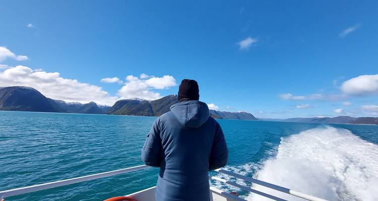 Traveler in jacket enjoys fast boat ride across bright turquoise fjord leaving foamy wake.