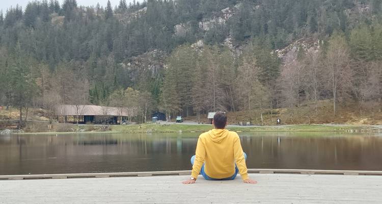 A person in a yellow hoodie sits on a wooden dock looking across a calm lake toward forested hills.