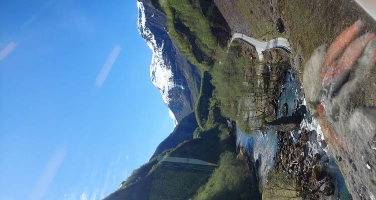 Mountain valley with winding river and snow-covered peak under a clear blue sky, photographed from above a road.