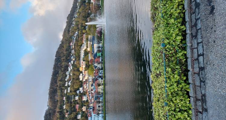 City lake with bubbling fountain, colorful hillside houses and a partly cloudy sky.