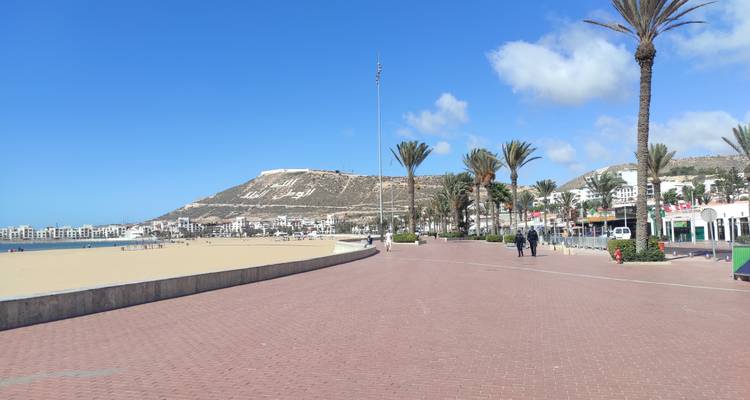 Large promenade en bord de plage avec le panneau d'Agadir sur la colline et des ciels bleus.