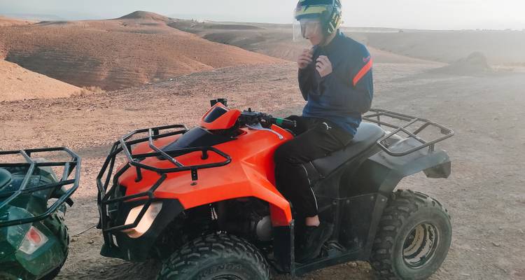 Un cavalier portant un casque est assis sur un quad orange sur un terrain désertique poussiéreux.