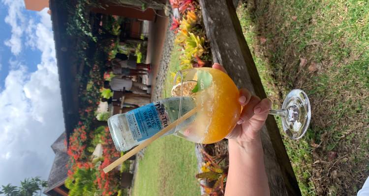 A hand holds a vibrant tropical cocktail garnished with an upside-down tonic bottle in a garden bar.