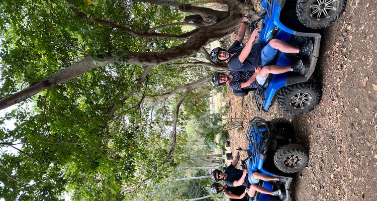 Four riders sit on blue quad bikes lined up beneath leafy trees during an off-road adventure.