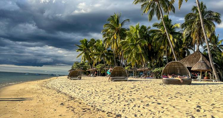 A sandy palm-lined beach dotted with woven loungers as dark dramatic clouds build over the turquoise sea.