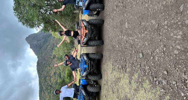 Five friends pose with raised arms beside their quad bikes against a rugged mountain backdrop.