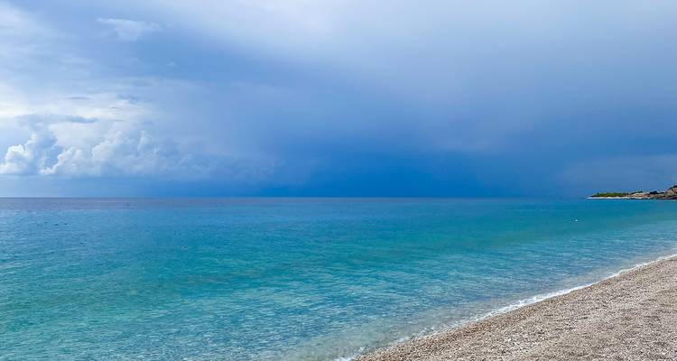 Mar turquesa tranquilo encontrándose con una playa de guijarros bajo un cielo de mal humor.