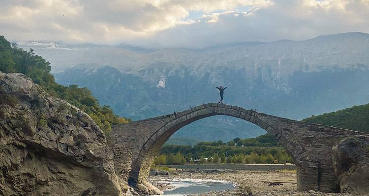Un aventurero de pie triunfalmente encima de un puente de arco de piedra antigua sobre un río rocoso.