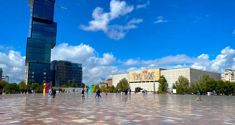 Plaza amplia en Tirana con rascacielos moderno, mural del Museo Nacional y peatones dispersos.