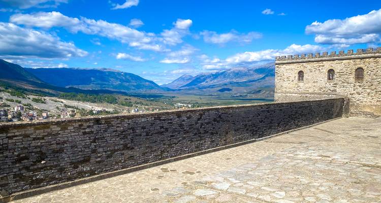 Les murs du château en pierre surplombent une large vallée et des montagnes bleues lointaines sous un ciel lumineux parsemé de nuages épars.