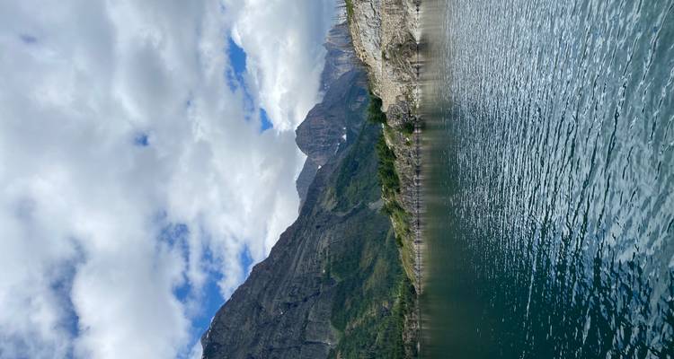 Ruhiger Bergsee mit kräuselnd bewegtem Wasser, bewaldete Hänge und felsige Gipfel unter teilweise bewölktem Himmel.