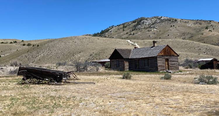 Historische Holzhütte und Wagen in einem trockenen Wiesental mit sanften Hügeln unter klarem blauen Himmel.