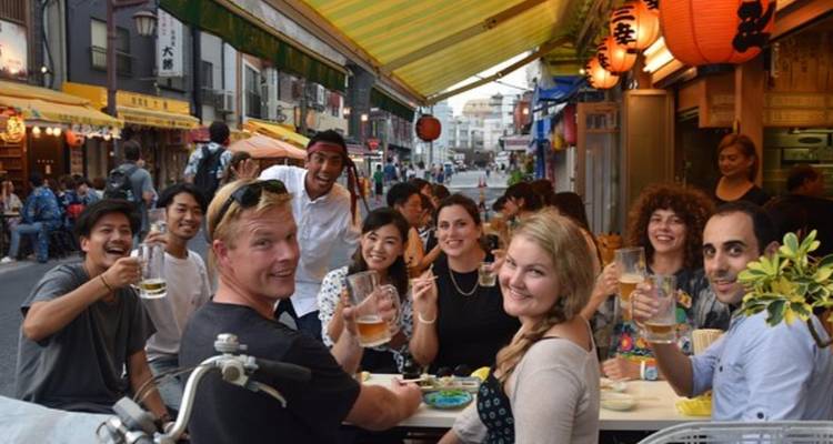 Lachende Gruppe von Reisenden, die Getränke in einer Freiluft-Izakaya-Straße in Tokio teilen.