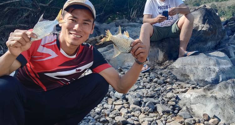 Homme souriant tenant fièrement deux poissons fraîchement pêchés tout en étant assis sur la rive rocheuse d'une rivière ; une autre personne utilise un téléphone derrière lui.