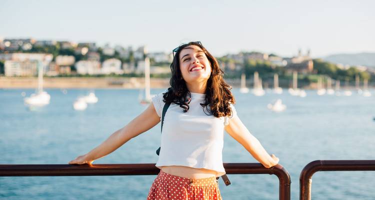 Smiling young woman leans against a railing with a sparkling bay full of sailboats and a hillside town behind her.