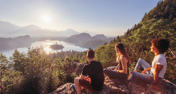Three friends sit on a rocky lookout above Lake Bled, watching the sunrise over the lake and surrounding mountains.