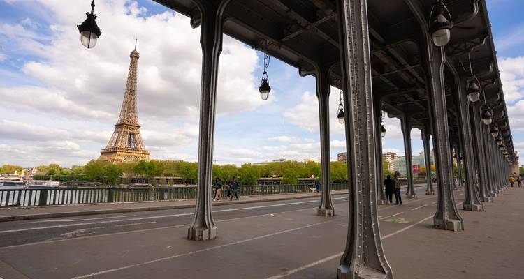 The Eiffel Tower rises beyond the iron columns of Pont de Bir-Hakeim with pedestrians on the bridge and the Seine below.