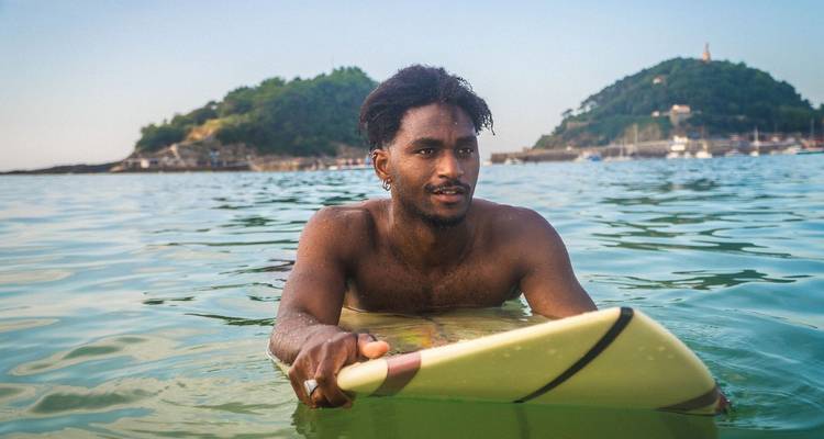 A surfer rests on his board in calm turquoise water with green islands and boats in the background.
