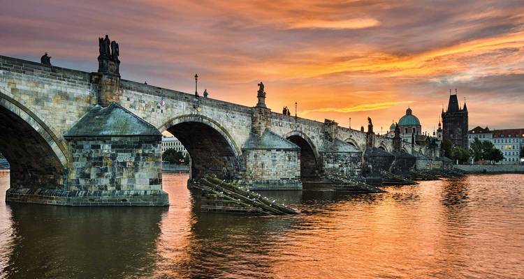 Charles Bridge spans the Vltava River under a glowing orange sunset sky in Prague.