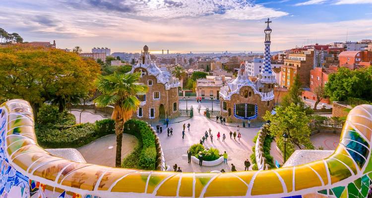 Colorful mosaic terrace of Park Güell overlooks Barcelona at sunrise with visitors in the plaza below.