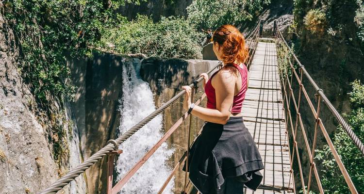 A woman pauses on a narrow suspension bridge overlooking a roaring gorge waterfall.