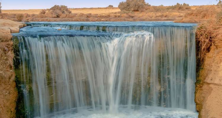 Une cascade aux eaux douces s'écoule dans une petite vasque entourée de sables désertiques et d'une végétation clairsemée.