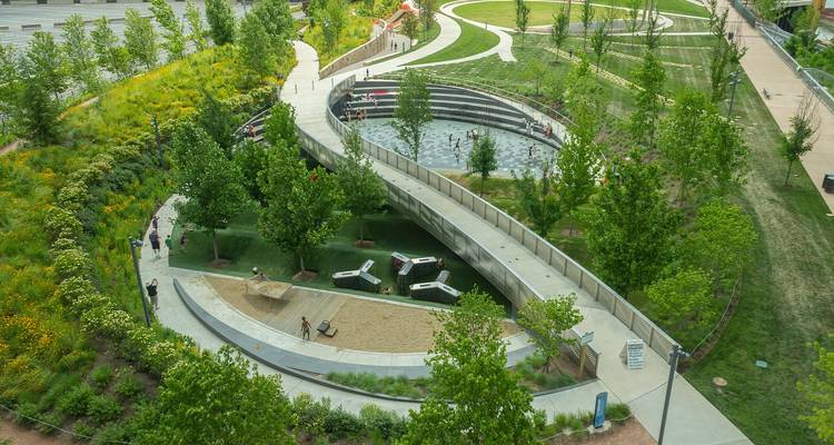 Vista aérea de un parque urbano moderno con caminos curvados, vegetación exuberante y visitantes paseándose alrededor de un área de fuente tipo anfiteatro.